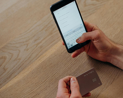 A man holding a phone in his right hand entering payment information, and a credit card in his left hand, over an oak table